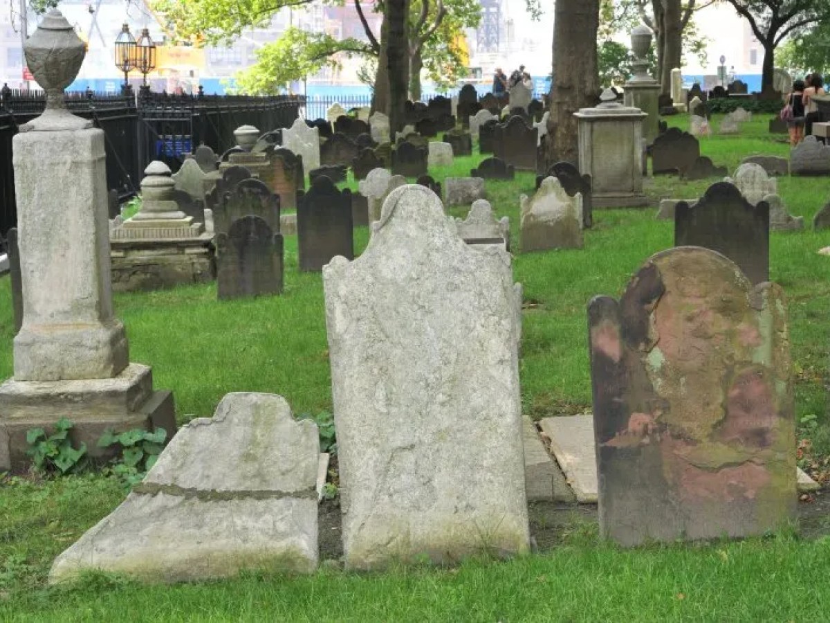 Old gravestones in a green cemetery with trees and a black fence.