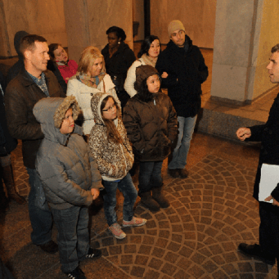 A man speaks to a group of people, including children, in a dimly lit indoor area.