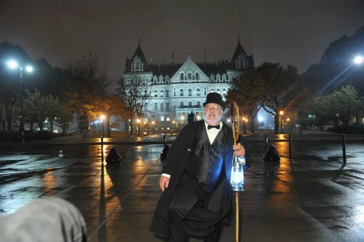 Man in vintage costume with lantern in front of illuminated historic building at night.