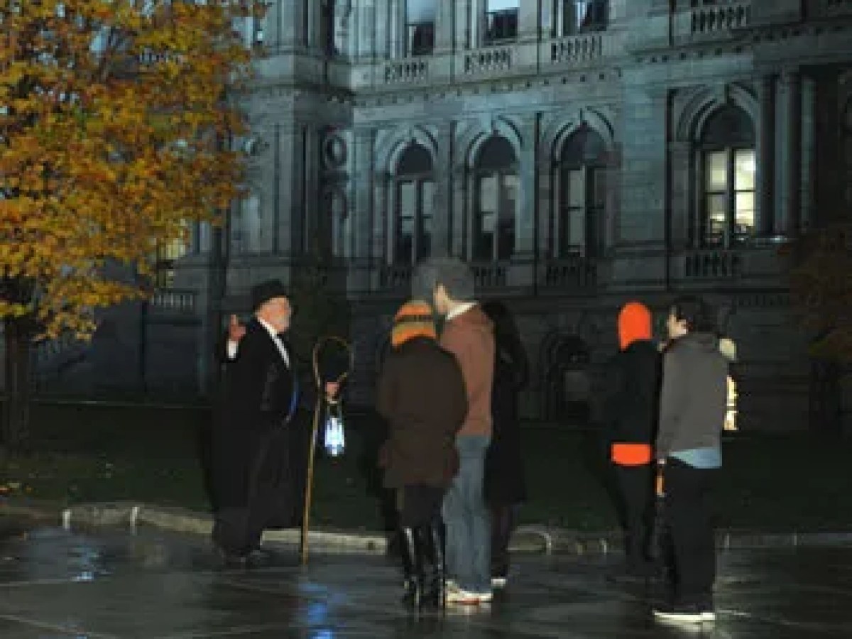 People gathered at night near a historic building with lit windows and autumn trees.