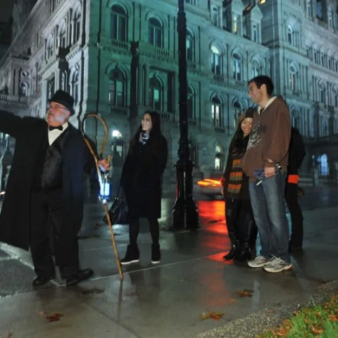 Tour guide points out a building to a group of people at night on a rainy street.