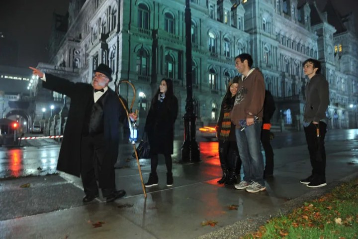 Tour guide points out a building to a group of people at night on a rainy street.
