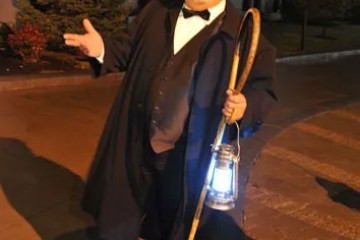 Man in 19th-century attire holding a lantern and cane, standing on a nighttime street.