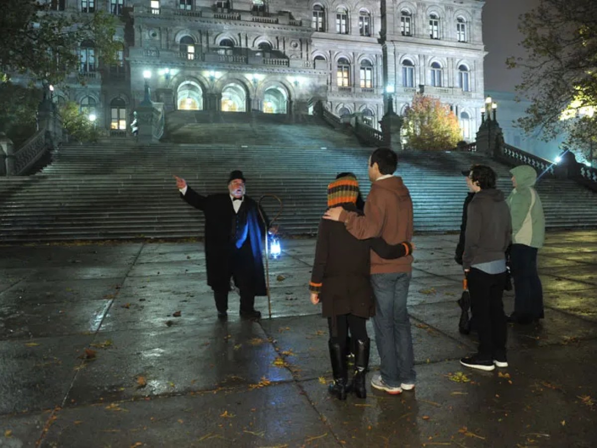 Tour guide with lantern speaks to group near illuminated grand staircase at night.