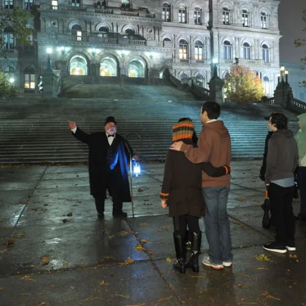 Tour guide with lantern speaks to group near illuminated grand staircase at night.
