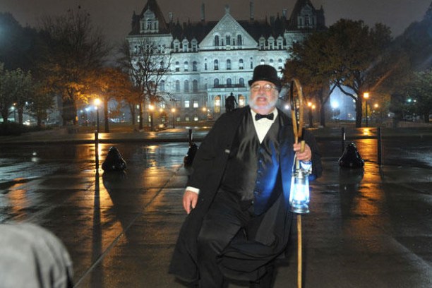 Man in formal attire and top hat holds lantern, standing on wet pavement with illuminated building behind.