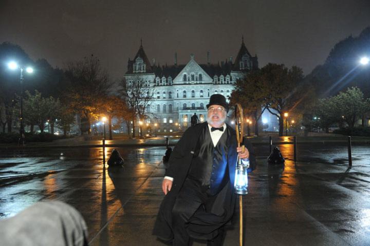 Man in period costume with lantern in front of large illuminated building at night.