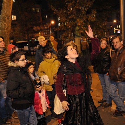 Group of people listening to a woman gesturing upwards on a nighttime city street.