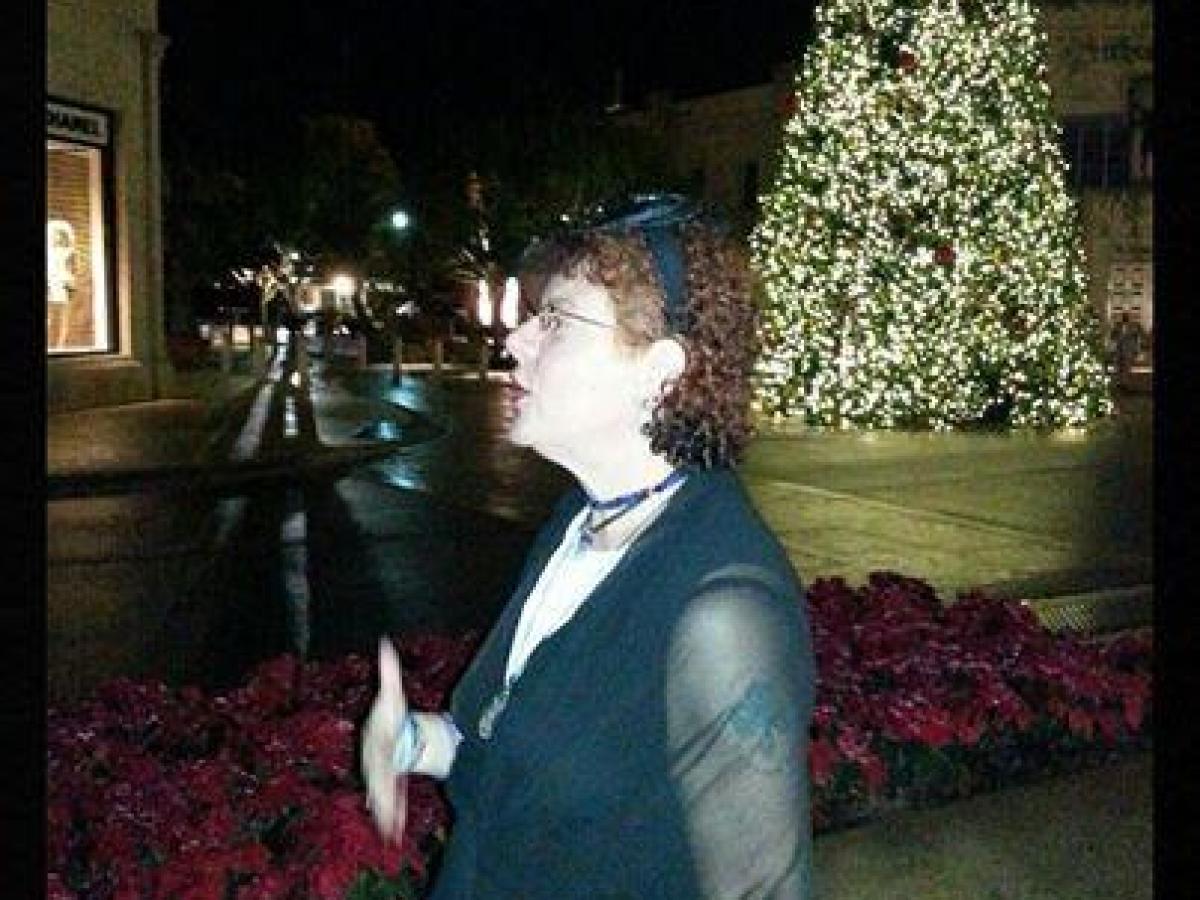 Woman speaking at night with a large lit Christmas tree and festive decorations in the background.