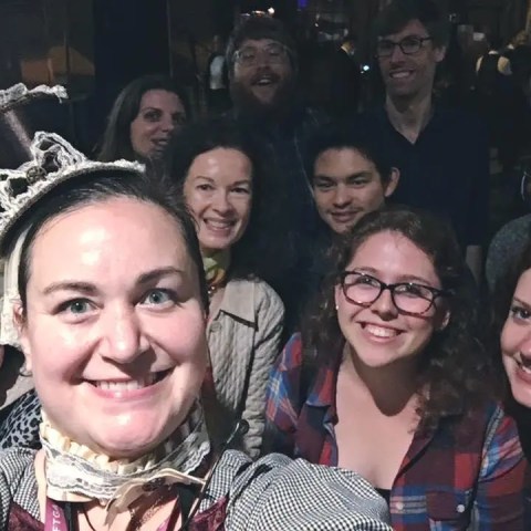 Group selfie with nine smiling people, one wearing a costume with a decorative headpiece.